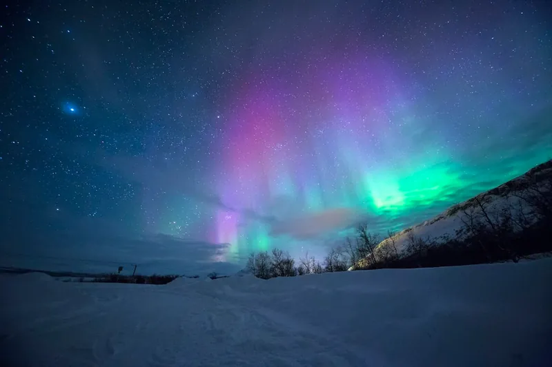 Cielo nocturno polar teñido de cortinas ondulantes de luz verde esmeralda y violeta sobre un lago congelado.